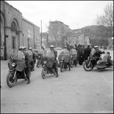 Immagine di un gruppo di 8 motociclisti e un sidecar dei Vigili Urbani in piazza Malatesta a lato del teatro Poletti. Il plotone di Vigili schierato e, sullo sfondo Castel Sigismondo con la casa del custode
